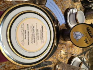 A table setting with a menu on a decorative plate, surrounded by teacups and glasses. The menu features meal options for a special event, and a "SHAKER the '66" sign is placed nearby on the golden tablecloth.