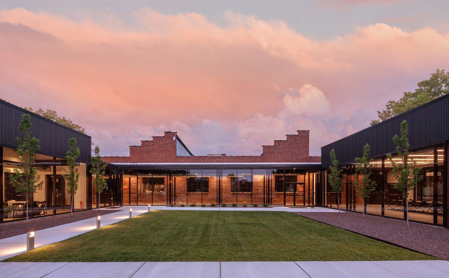 Modern brick building with glass elements under a pink sky. Green lawn and pathway in the foreground.