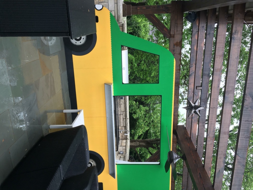 A green and yellow food truck is parked under a wooden pergola. The truck has no visible sign or logo and is surrounded by greenery. Nearby, dark wicker furniture is arranged on a tiled patio.