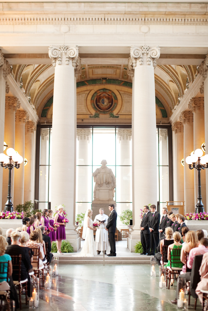 A wedding ceremony inside a grand, classical building with high ceilings and large pillars. The couple stands before an officiant, surrounded by their bridal party dressed in purple. Guests are seated, and the aisle is lined with candles.