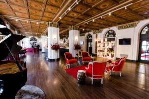 Elegant hotel lobby with red velvet chairs and sofas, dark wood flooring, and ornate wooden ceiling. A grand piano is in the corner, and a bookshelf lines the wall. Large floral arrangements and circular tables add to the luxurious atmosphere.