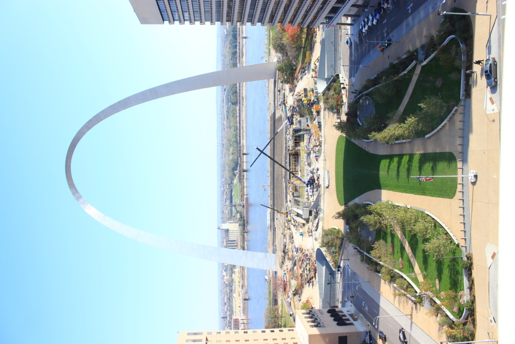 Aerial view of the Gateway Arch in St. Louis, with the Mississippi River in the background. The surrounding area features a green park and pathways, flanked by tall buildings.