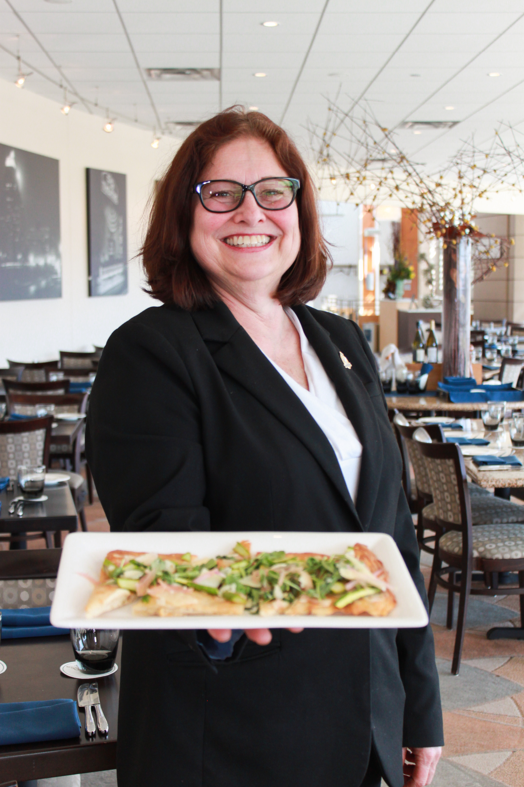 A smiling woman in a black suit holds a rectangular plate with a dish garnished with greens. She stands in a well-lit restaurant with neatly arranged tables and modern decor in the background.