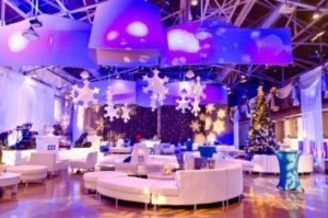 A festive indoor setting with purple and blue lighting, featuring large hanging snowflakes, white couches, and a decorated Christmas tree. The room has a modern design with high ceilings and a well-lit, elegant atmosphere.