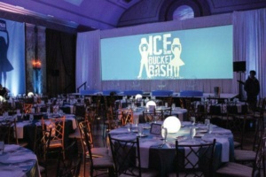 A decorated event space with round tables set for guests. A large screen displays "Ice Bucket Bash" with silhouetted figures. The lighting is dim with glowing centerpieces illuminating the tables.