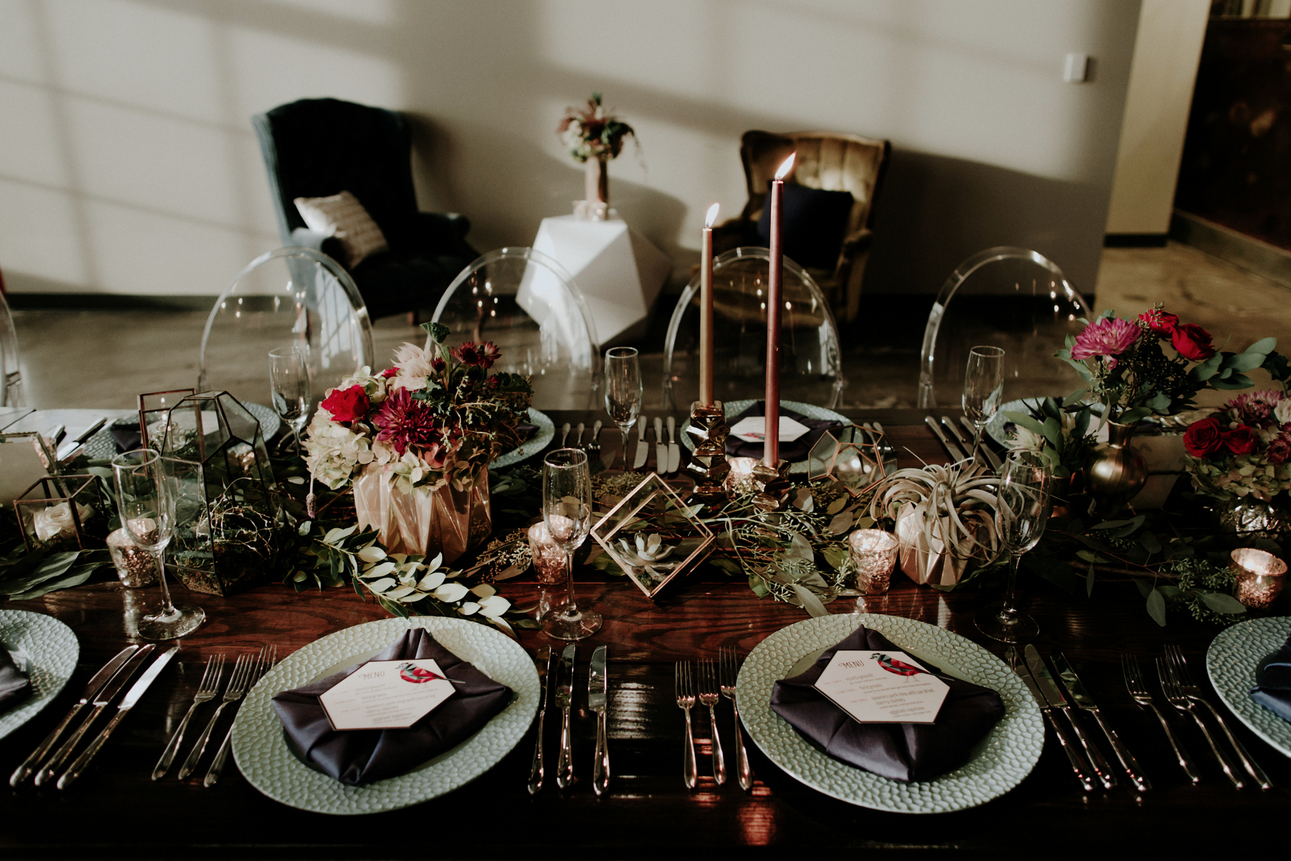 A beautifully set dining table with elegant floral arrangements, candles, and geometric decor. Plates are set with dark napkins and menus. Sunlight casts soft shadows across the table, creating a warm and inviting atmosphere.