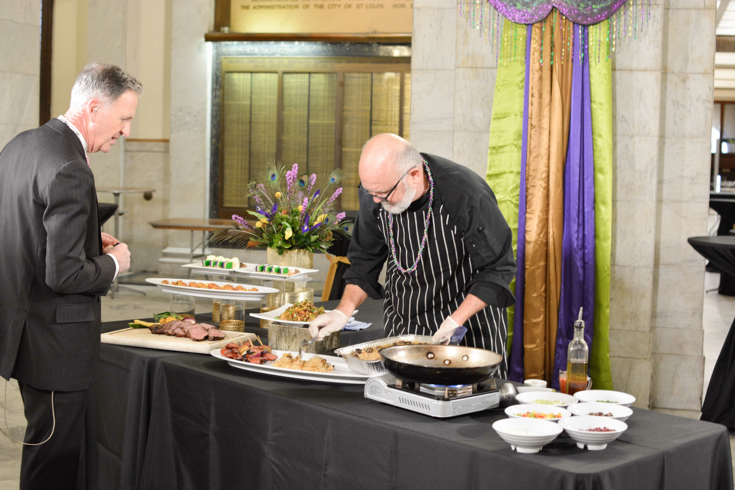 A chef in a black uniform prepares dishes at a food station adorned with colorful drapery and floral arrangements. A man in a suit observes the presentation. Various plated foods and bowls are displayed on the table.