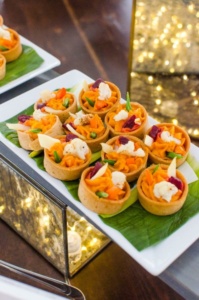 A platter of colorful savory tarts filled with assorted vegetables, including carrots, cauliflower, and greens, is placed on a large leaf. The background features decorative fairy lights creating a warm ambiance.