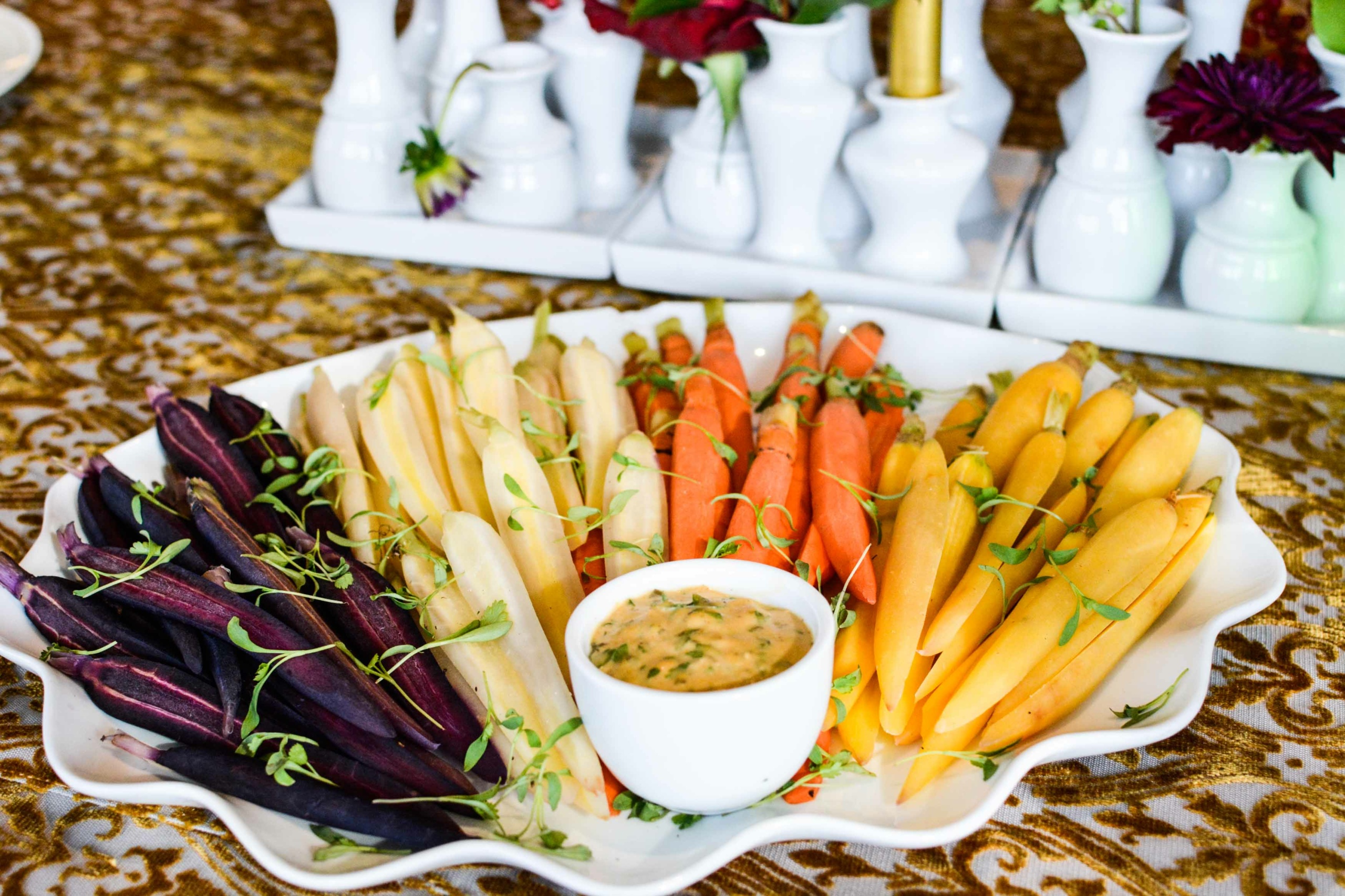 A platter of assorted colorful carrots—purple, white, orange, and yellow—arranged in sections around a bowl of dipping sauce, garnished with small greens. The platter is set on a gold-patterned tablecloth with white vases in the background.