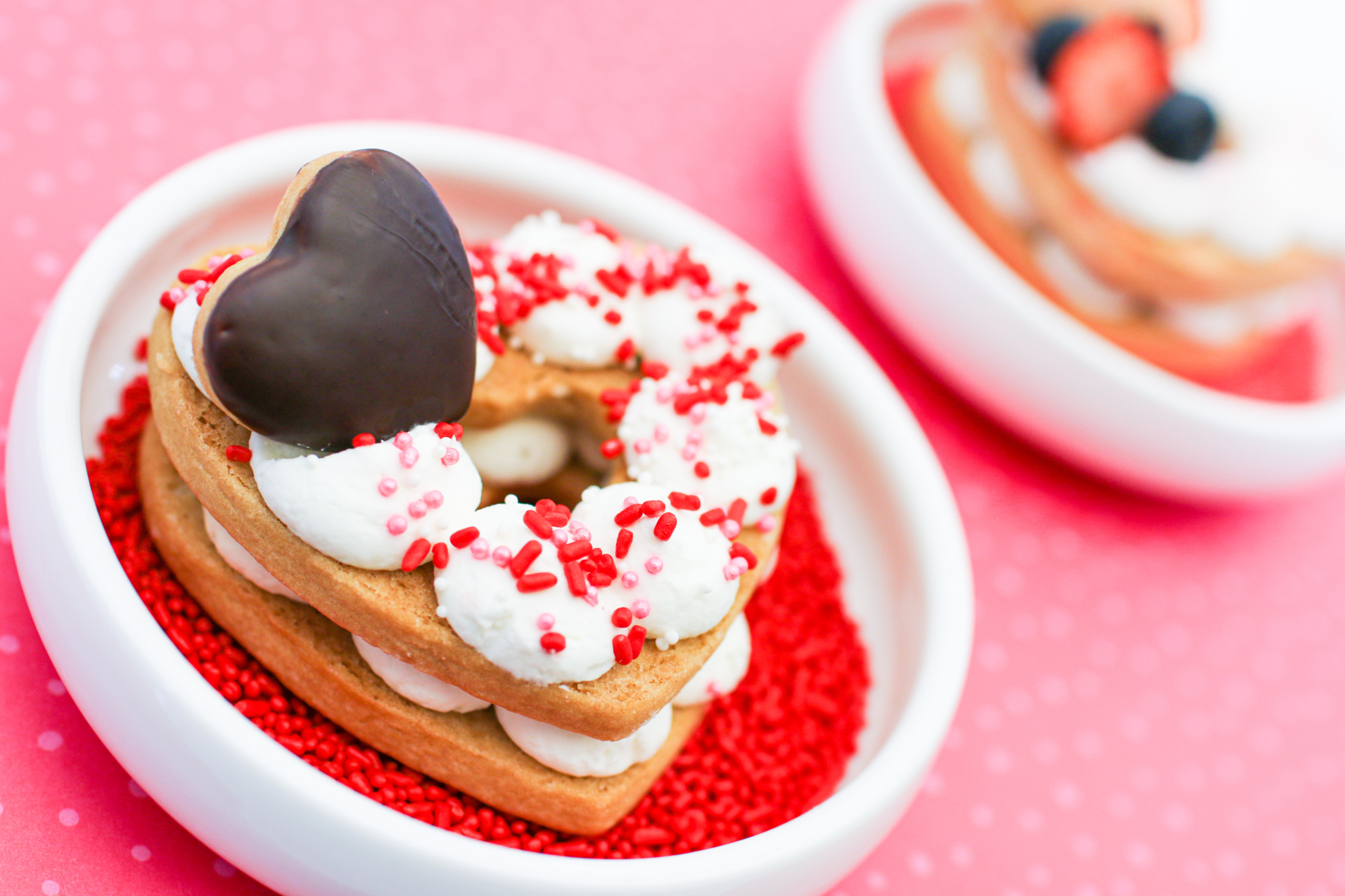 Heart-shaped dessert topped with white frosting, red sprinkles, and a chocolate heart sits in a bowl with red sprinkles. Another similar dessert is partially visible in the background, garnished with strawberries and blueberries.