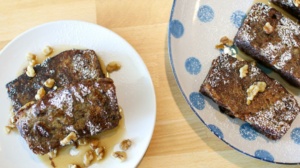 Two plates of French toast topped with powdered sugar and walnuts. One plate is white, holding two slices, and the other is blue with polka dots, holding three slices. Both plates are on a wooden table.