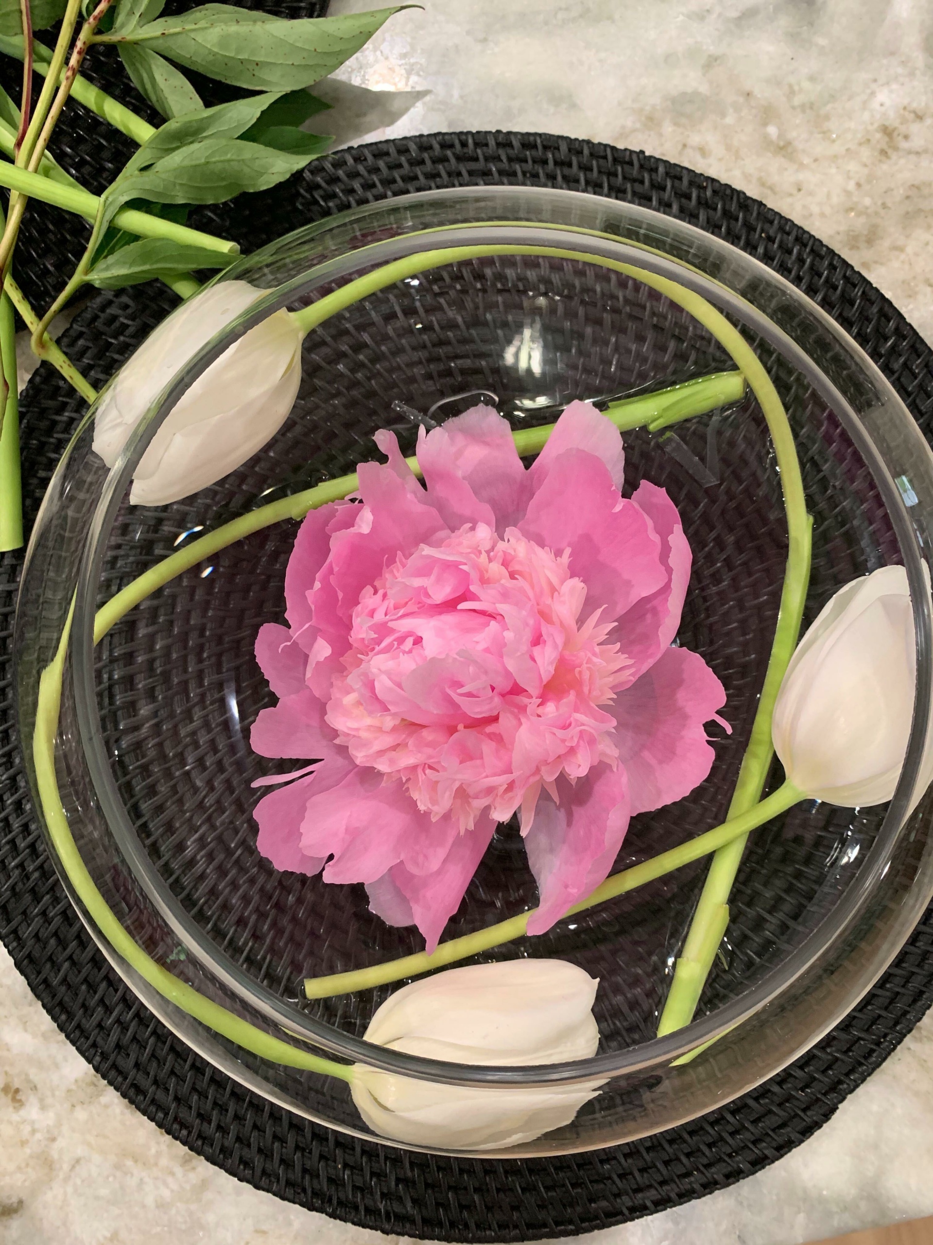 A pink peony floats in a clear glass bowl of water, surrounded by white tulip buds. The bowl rests on a black woven placemat, creating a contrasting background. Green leaves are visible in the top left corner.