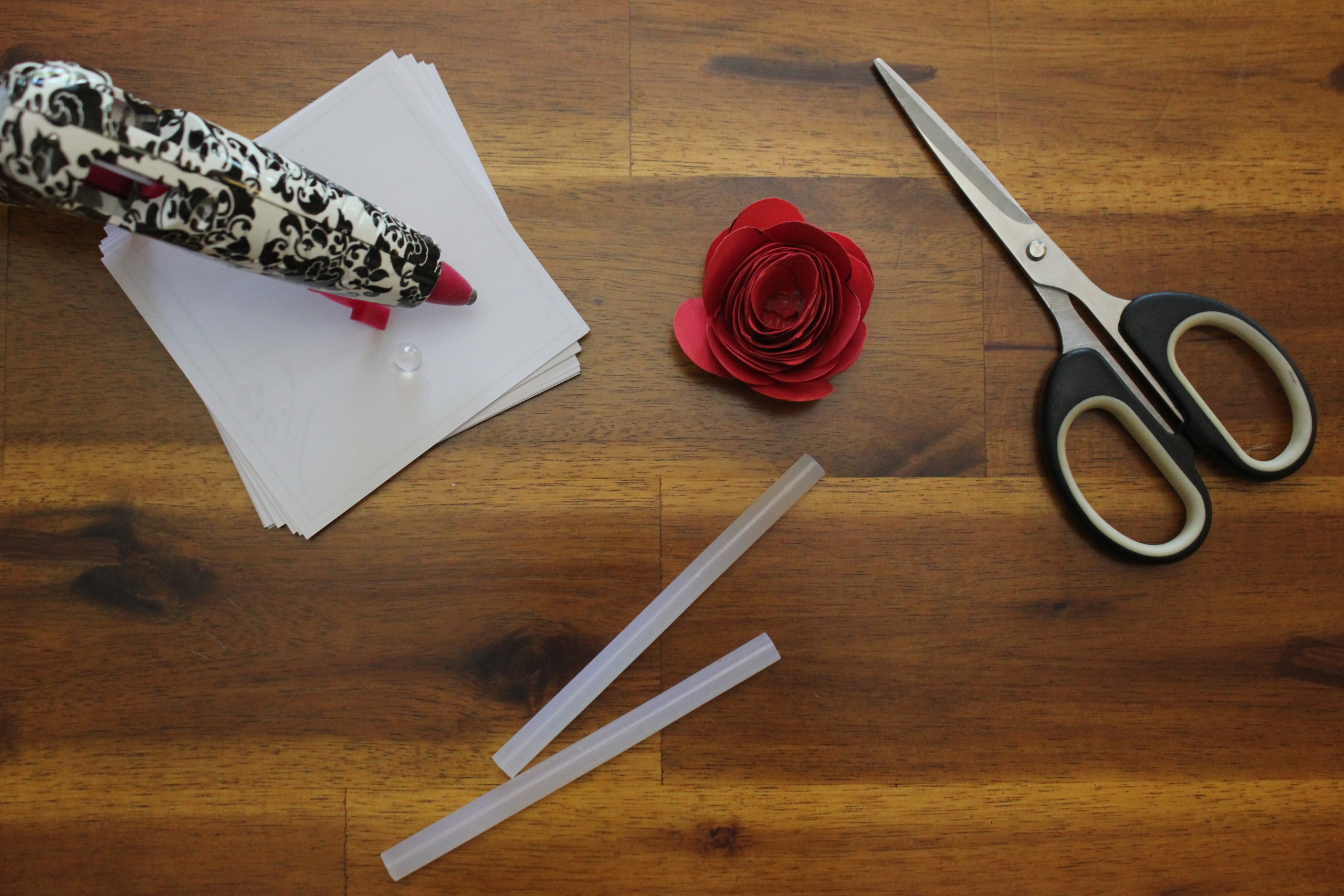 A DIY crafting setup on a wooden table featuring a glue gun, stack of white paper, red paper flower, scissors, and glue sticks.