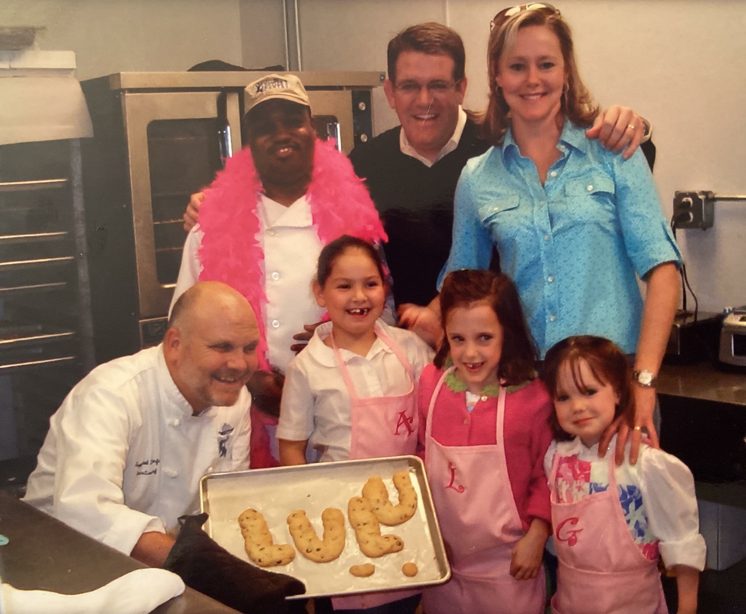 A group of adults and children in a kitchen, smiling. One adult holds a tray of cookies shaped like letters. Some are wearing aprons and one wears a pink feather boa. There is a commercial oven in the background.