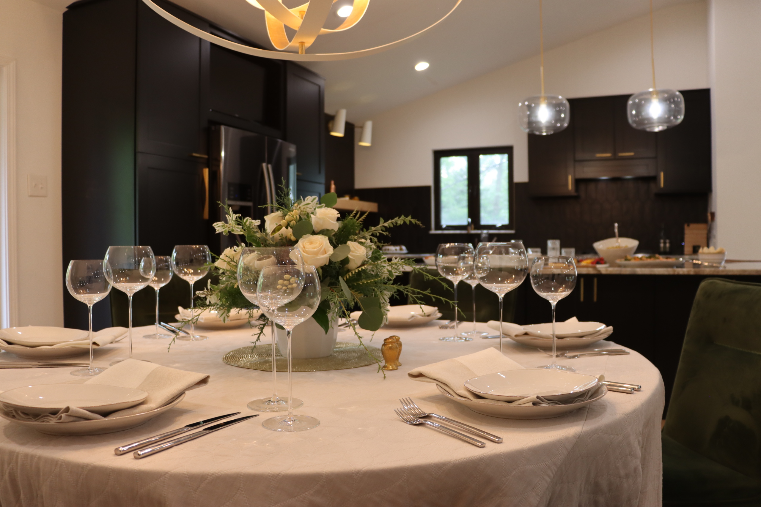 A dining table set for six with elegant glassware, white napkins, and a floral centerpiece of white roses. The background features a modern kitchen with dark cabinets and pendant lights.