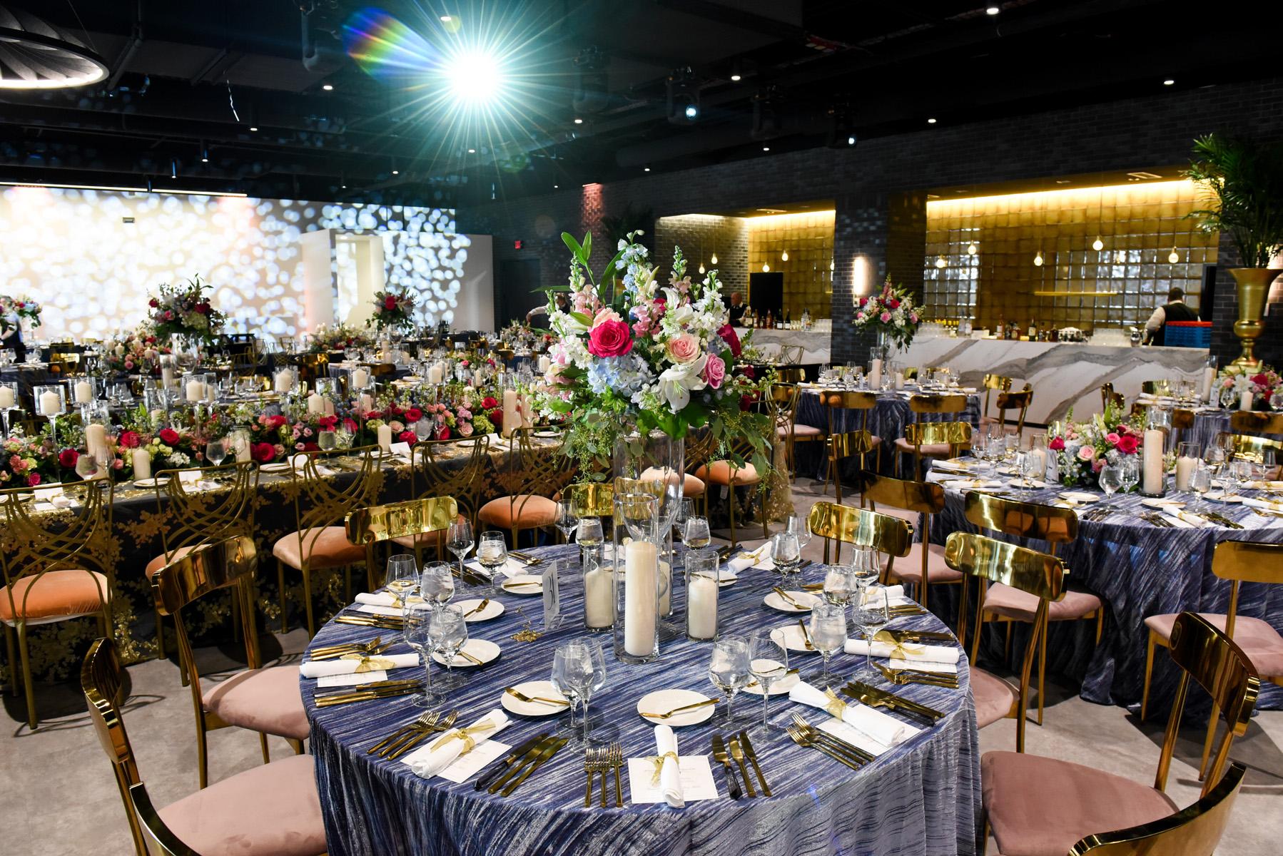 A lavish event setup featuring round tables draped with blue linens, adorned with floral centerpieces and white candles. The room is decorated with elegant lighting and gold chairs, creating a sophisticated atmosphere.