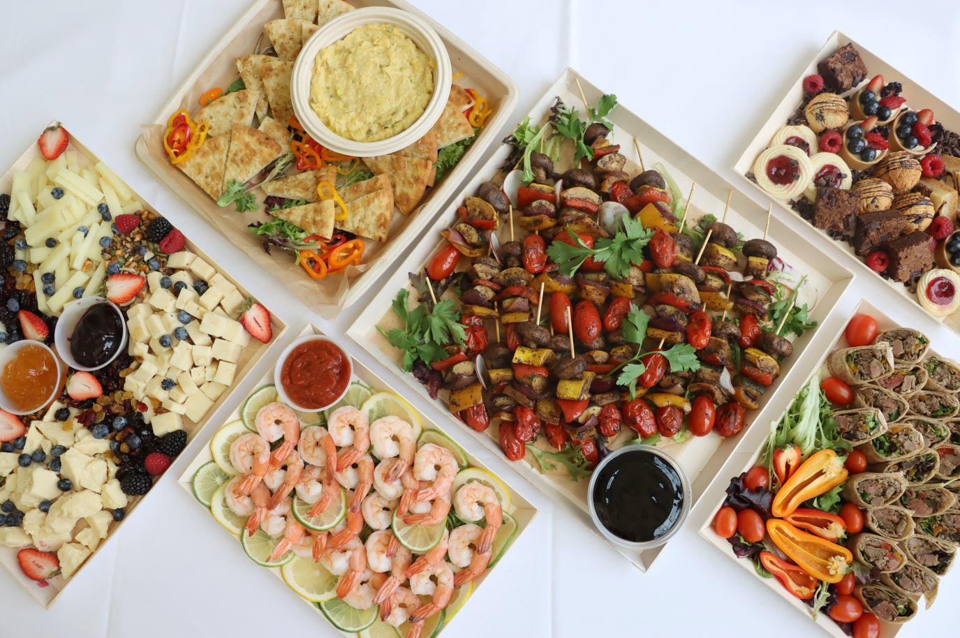 An assortment of platters on a table, featuring cheese and fruit, shrimp with lime slices, vegetable skewers, filled pastries, brownies, and a platter with dip and pita bread. Garnishes of parsley and bell peppers are scattered around.