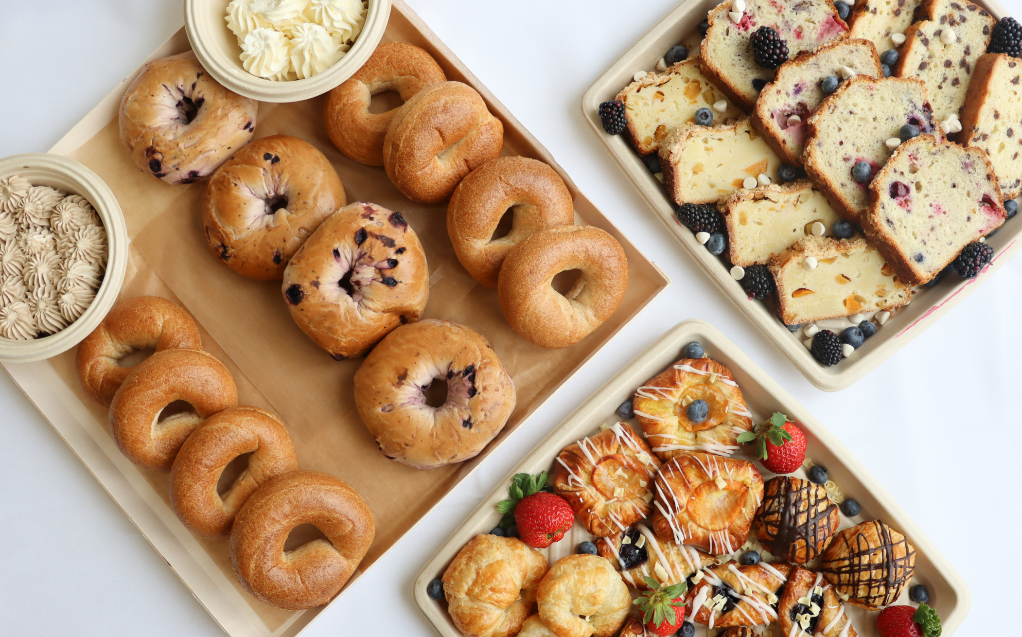 Three trays on a table: one with assorted bagels and cream cheese, another with various slices of fruit-studded loaf cakes, and the last with an assortment of pastries, including croissants, turnovers, and decorated danishes, garnished with strawberries.