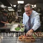 A man in a professional kitchen arranges salad leaves on a tray. He wears glasses, a white shirt, and a bright tie. Behind him, another person is preparing food. The kitchen is bustling with stainless steel equipment. Text overlays the image.