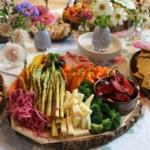 A colorful platter featuring asparagus, cheese, broccoli, sliced plums, carrots, mini bell peppers, and pickled onions on a wooden base. Surrounded by bowls of crackers, bread, and small vases of flowers on a table.