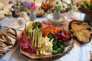 A colorful platter featuring asparagus, cheese, broccoli, sliced plums, carrots, mini bell peppers, and pickled onions on a wooden base. Surrounded by bowls of crackers, bread, and small vases of flowers on a table.