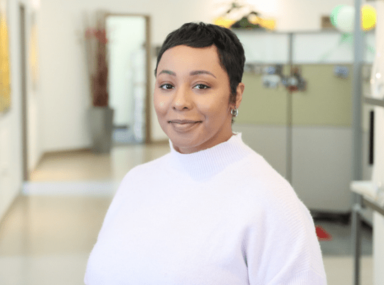 Person with short hair smiling in an office setting, wearing a light sweater.