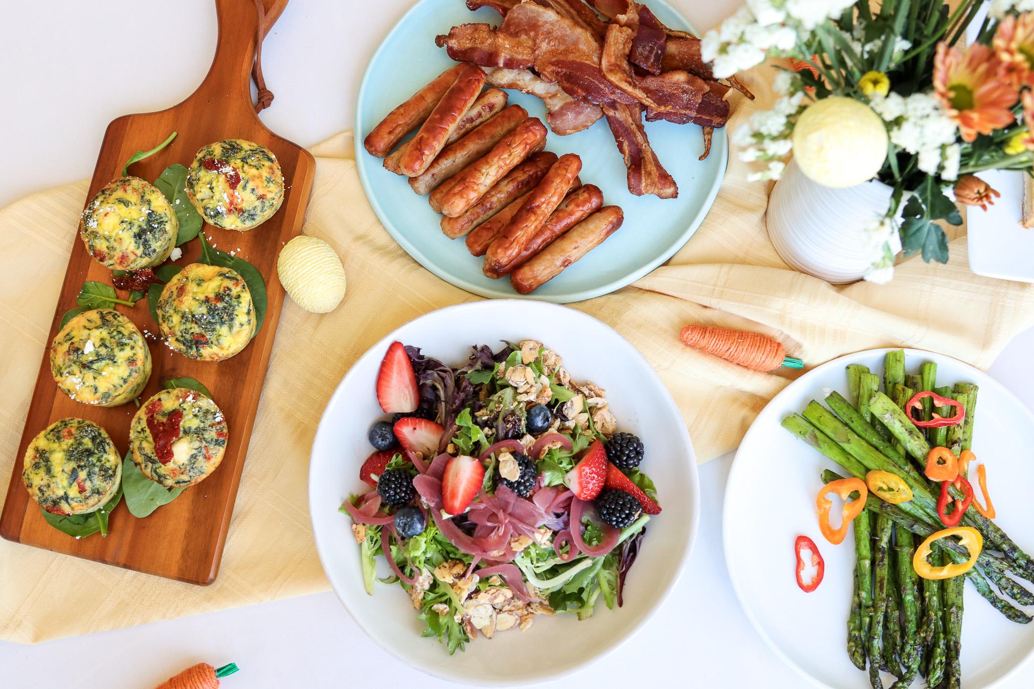 A table with a salad, asparagus, baked egg muffins, sausages, bacon, and flowers for decoration.