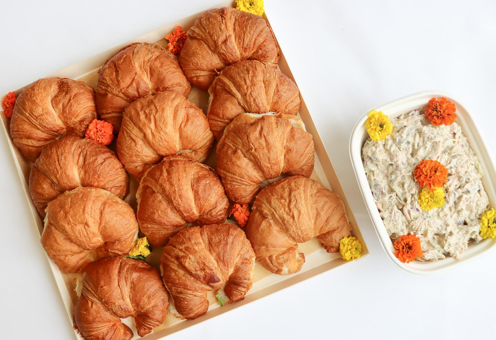 A tray of croissant sandwiches alongside a bowl of salad, garnished with colorful flowers on a white background.