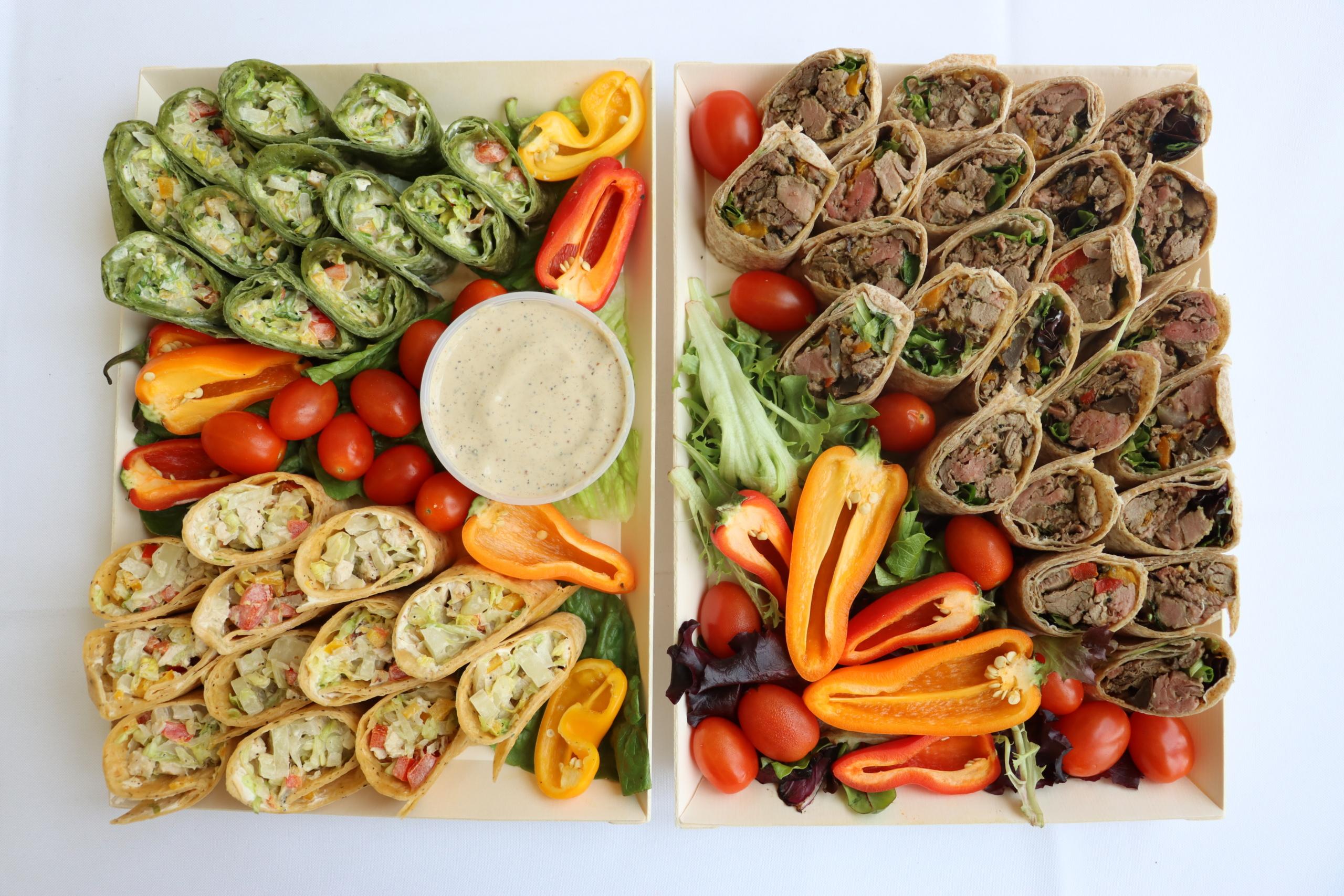 Two trays of wrap sandwiches with dip, assorted bell peppers, and cherry tomatoes.