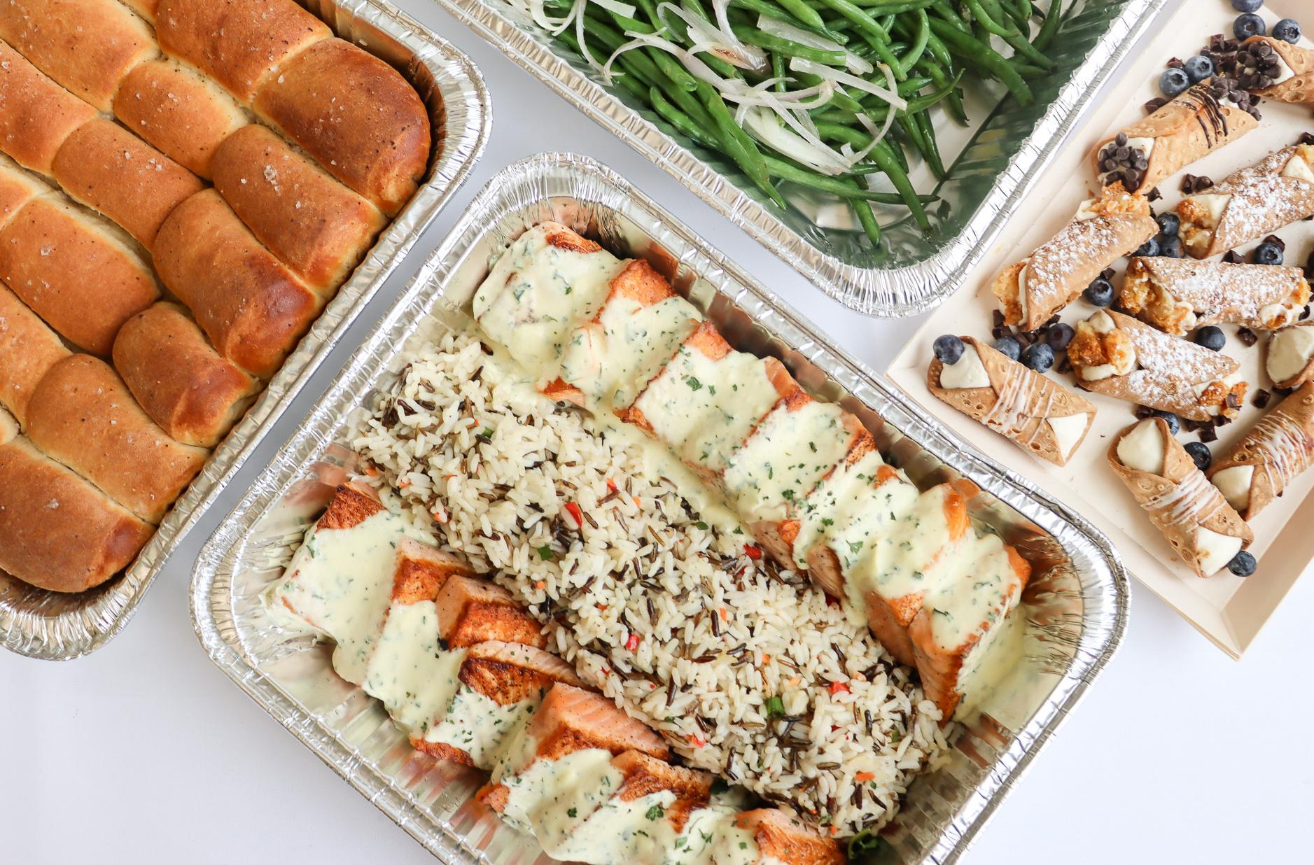 Catering trays with bread rolls, green beans, salmon with rice, and dessert cannoli.