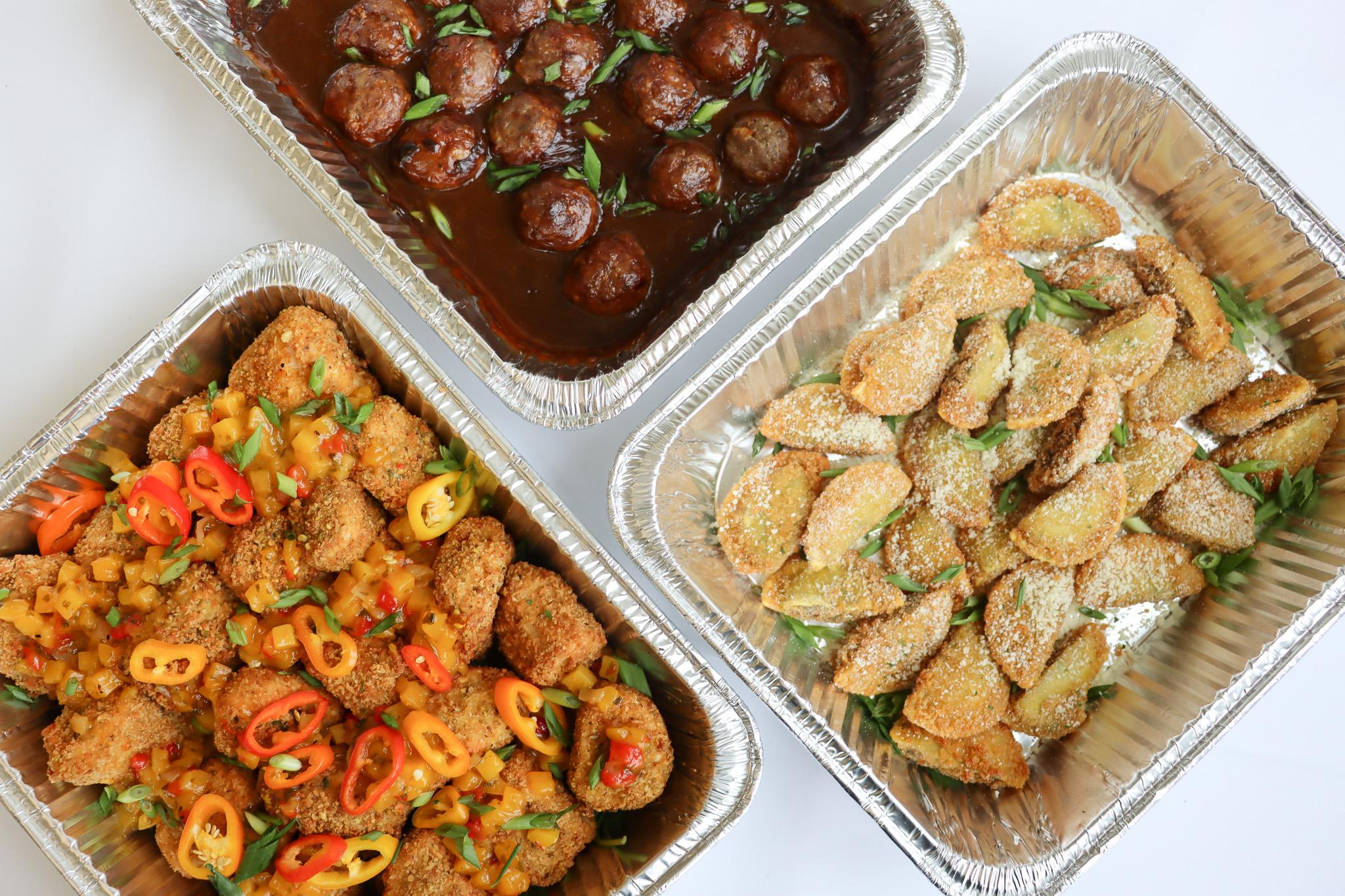 Three foil trays with meatballs in sauce, breaded chicken with peppers, and seasoned potato wedges.