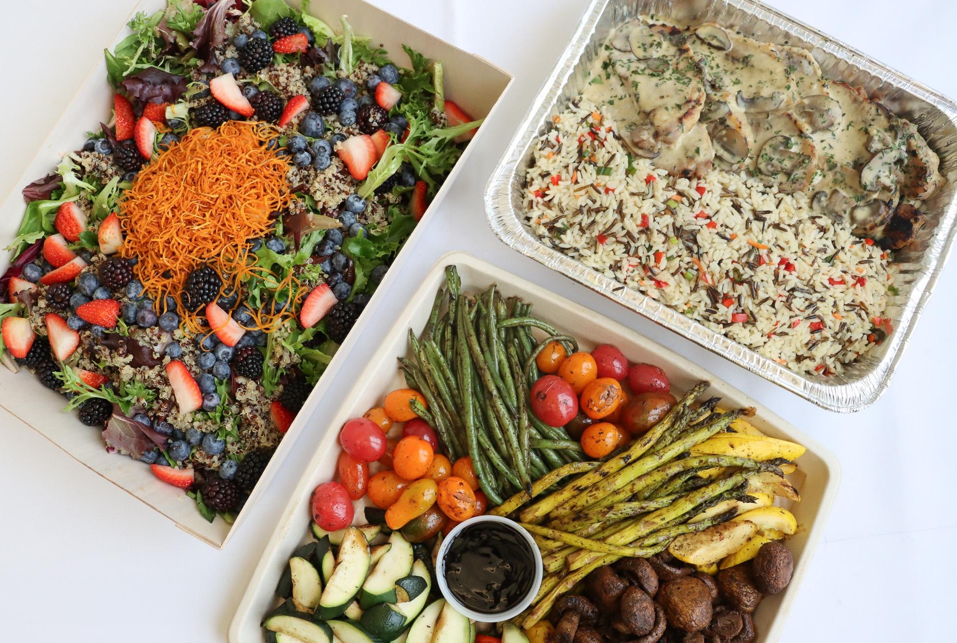 Three trays of food: mixed berry salad, herb rice with sauce, and assorted grilled vegetables with a dipping sauce.